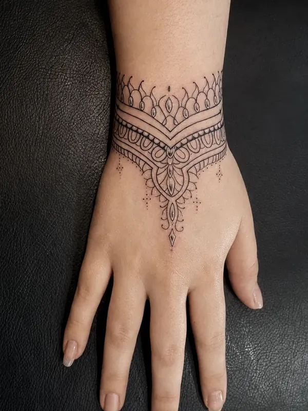 Tatuagem fine line ornamental em bracelete no punho, com pingente descendo pelo dorso da mão