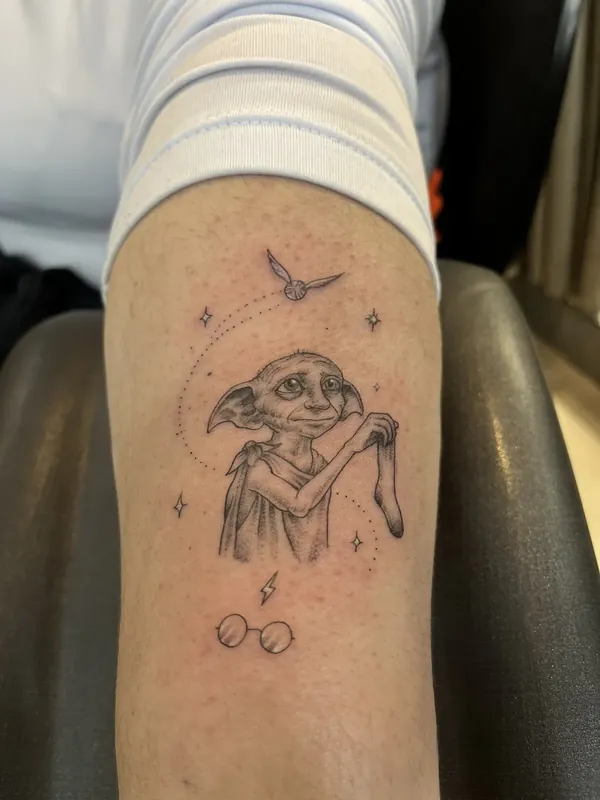 Tatuagem fine line do elfo Dobby segurando a meia, com óculos redondos e um pomo, no braço