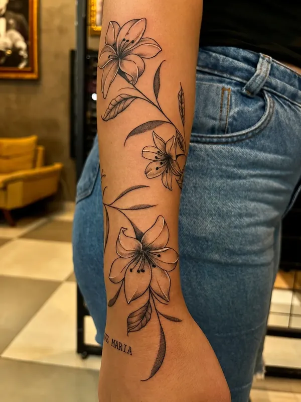Tatuagem fine line de lírios subindo pelo braço, com a frase Ave Maria junto ao punho
